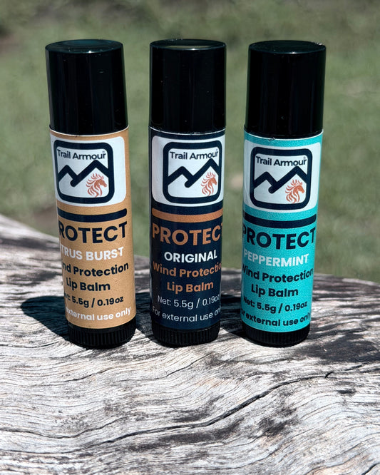 Three Trail Armour PROTECT lip balms standing on a weathered log: Citrus Burst, Original and Peppermint