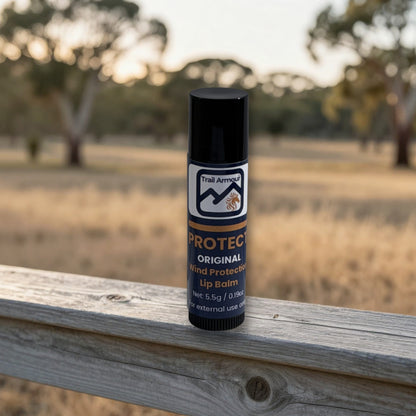 A single tube of Trail Armor Protect Original Wind Protected Lip Balm stands upright on a weathered wooden fence with a blurred outdoor field and trees in the background.