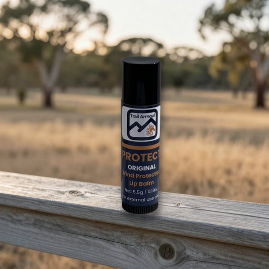 A single tube of Trail Armor Protect Original Wind Protected Lip Balm stands upright on a weathered wooden fence with a blurred outdoor field and trees in the background.